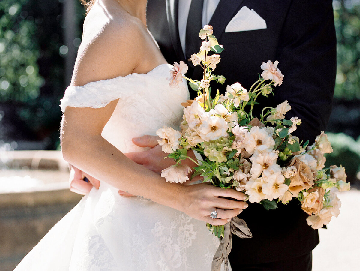 Close-up of a bride in a lace dress holding a peach and cream bouquet, arm in arm with a groom in a dark suit, soft natural light