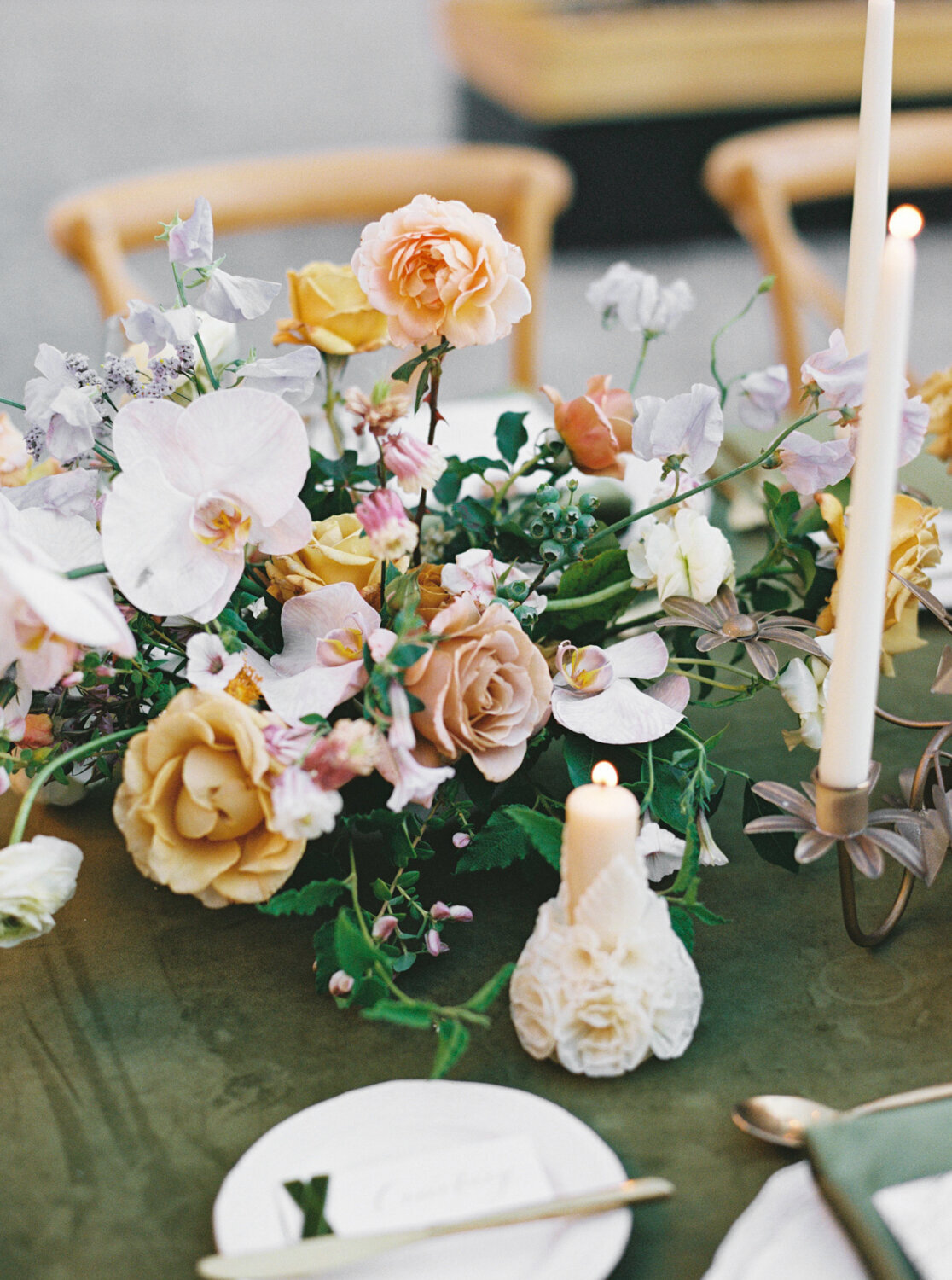 Close-up of a lush floral centerpiece with peach, yellow, and white flowers, greenery, and lit taper candles on a table