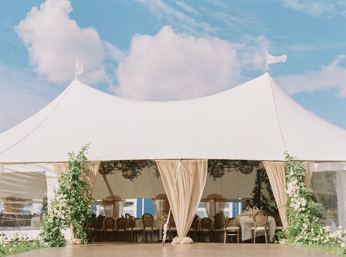 Large white event tent with draped curtains and floral arrangements, set against a blue sky with clouds, elegant outdoor setting